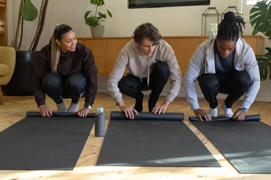Diverse friends unrolling black yoga mats in living room at home in athleisure with water bottle