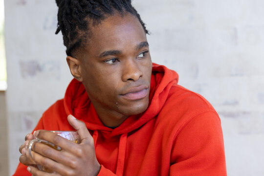 African man holding clear glass mug with both hands at table in red hoodie, gazing left