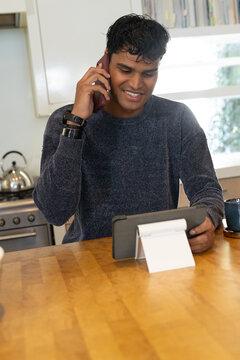 Indian man talking on smartphone while checking tablet at wood kitchen counter wearing dark sweater