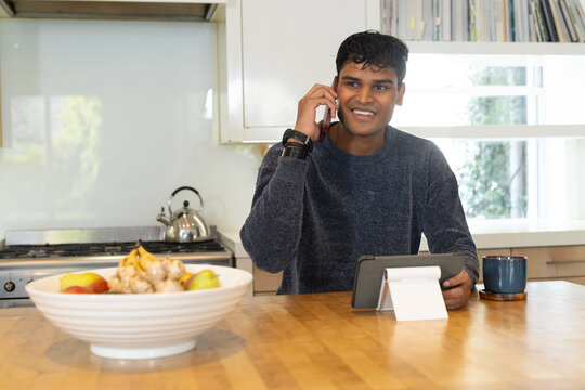 Adult Indian man speaking on smartphone, steadying tablet on white stand at kitchen table