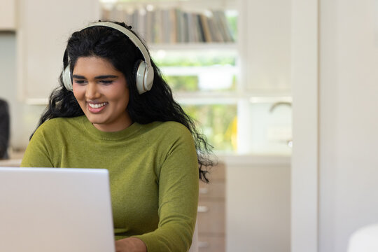 Indian woman sitting at kitchen nook, working on silver laptop wearing green sweater and headphones