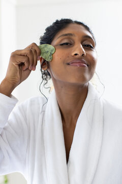 African American woman massaging face with green gua sha stone in bathroom wearing white bathrobe