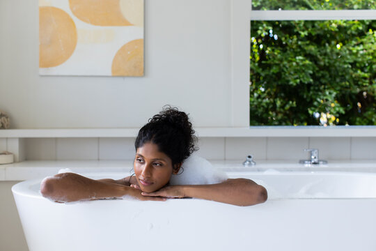 African American woman reclining in freestanding bathtub at home, surrounded by foam