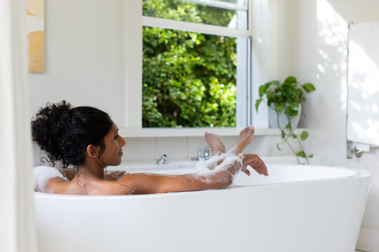 African American woman reclining in freestanding bathtub, resting on rim with foam and plant