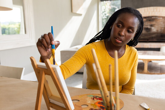 African American woman wearing mustard top painting on table easel at home with palette and brushes