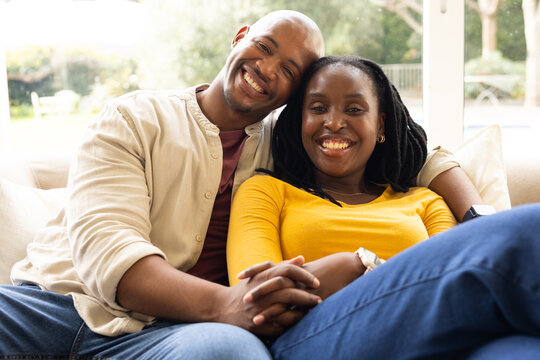 African couple sitting closely on light sofa by large window in living room, wearing jeans, smiling