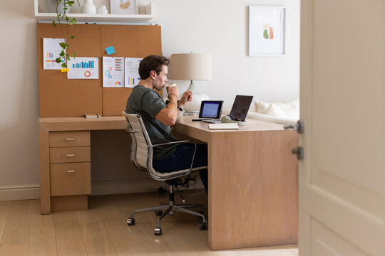 Man sipping from ceramic mug while working on laptop and tablet in home office checking corkboard