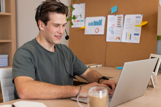 Man wearing short-sleeve t-shirt and headset with boom mic typing on laptop at desk with corkboard