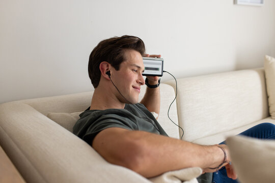 Man in his twenties reclining on beige sofa holding audio player against temple wearing earbuds