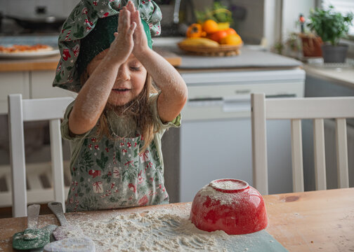 Cute little girl wearing chef hat claps hands covered in white flour over wooden table. Christmas home baking process perfect for cooking class or family activity themes