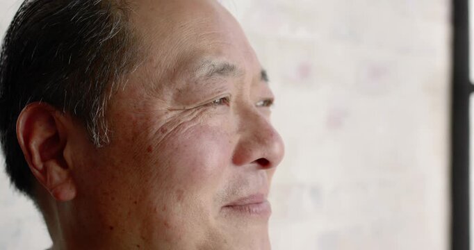 Senior Asian man smiling and laughing while responding to off-camera voice near bright window