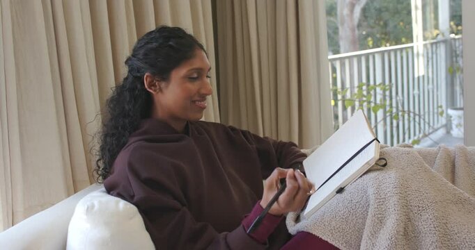 Woman sitting on sofa at home in hoodie writing notebook twirling pen reflecting gazing outside