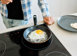 A man cooks a sunny-side-up egg in a small skillet on a modern electric stovetop while preparing a...
