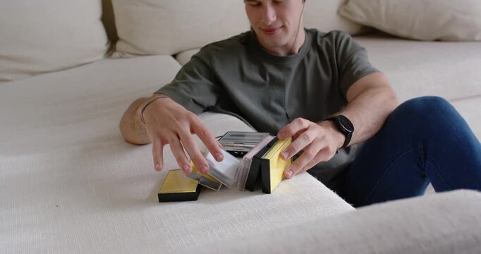 Man in mid-20s sitting on sofa noticing yellow-and-black boxes, opening and inspecting clear cases