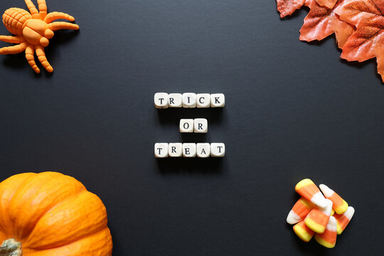 Trick or treat spelled with letter tiles on black background with pumpkin, candy corn, toy spider and autumn leaves.
