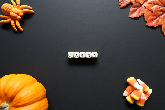Candy spelled with letter tiles on black background with pumpkin, candy corn, toy spider and autumn leaves.