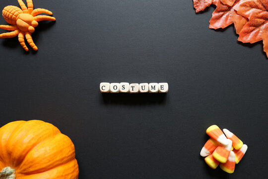 Costume spelled with letter tiles on black background with pumpkin, candy corn, toy spider and autumn leaves for a Halloween dress-up concept.