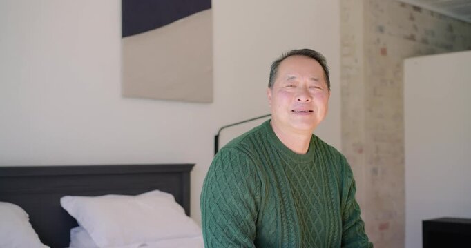 Asian senior man on bed in green-knit turning to cam, smiling-laughing on off-camera cue