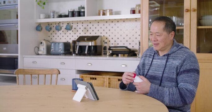 Mature Asian man being prompted by tablet holding white bottle in kitchen reporting throat pain