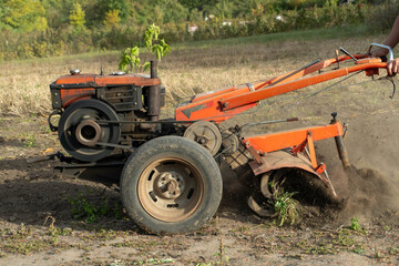 Farmer operates walk behind tractor to till dry field soil. Orange rotary cultivator churns earth and uproots weeds, raising dust as operator grips handles and guides machine across open rural plot. © Віталій Б.