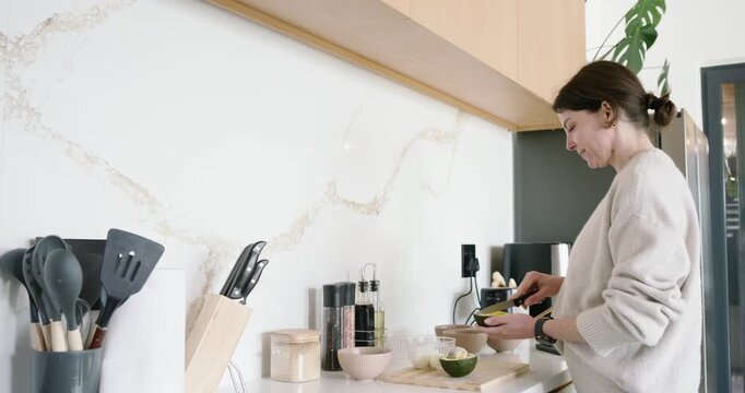 Woman in her 30s preparing breakfast, halving avocado, scooping into bowls, using toaster