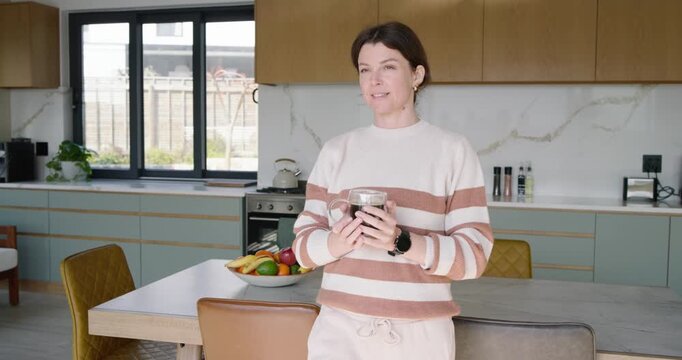 Woman leaning on kitchen island, morning light urging sipping glass mug while camera pushing in