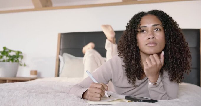 Adult woman lying on bed planning and writing with pen in notebook, pausing then tapping smartphone