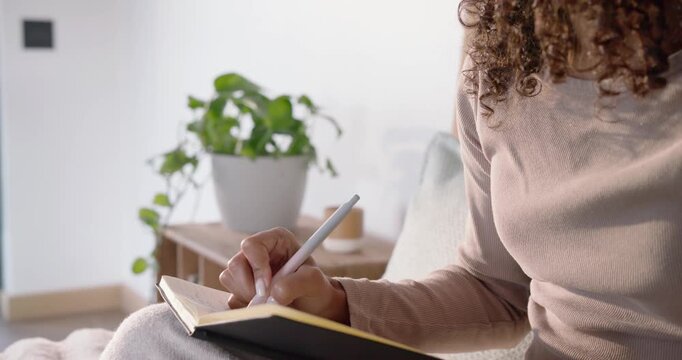 African American woman journaling with stylus in nook, pausing as daylight draws gaze, copy space