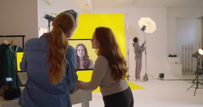 Two women at desk checking screen to refine pose, photog pointing, model miming