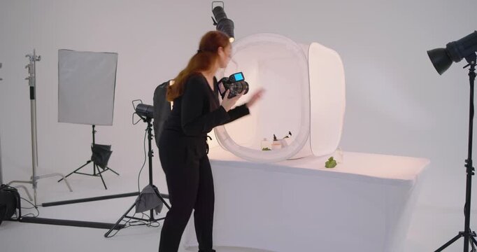 Entering studio woman shooter arranging props in light tent, testing DSLR flash for product shoot