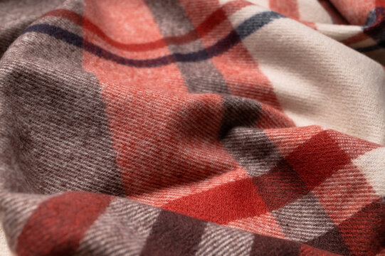 Warm plaid fabric forms soft waves in close view, showing red, beige and gray checks with tactile weave. Textile material suits scarf design, autumn clothing, cozy styling and retail use.