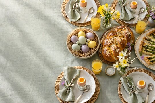 Easter brunch table from above with soft boiled eggs, braided sweet bread, pastel dyed eggs in nest, asparagus tart, juice and daffodils