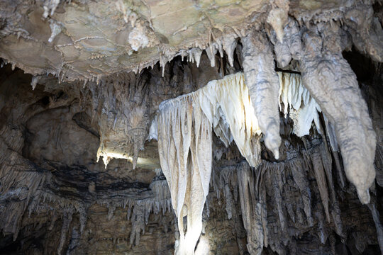 A wide perspective of a stunning subterranean ceiling featuring complex, curtain-like limestone draperies and sharp stalactites, softly lit against the dark rock background.