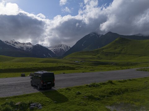 A van on a road in a green mountain landscape under a cloudy sky, aerial view, camper, motorhome, mountain landscape between Gudauri and the Cross Pass, Sherkhota Mountain, Mtskheta-Mtianeti region, Georgian Military Road, High Caucasus, Georgia