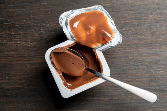 Chocolate pudding in plastic cup with spoon, closeup. Top view of opened white plastic container with sweet chocolate mousse or yogurt. Creamy dairy dessert with metal spoon on dark wooden table