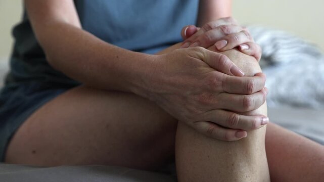 Female hands tightly clutching painful knee while trying to sit up in bedroom, muscle strain