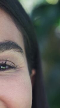 Vertical video: Reacting to sunlight, woman opening left eye and smiling under leaf bokeh outdoors