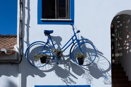 Blue bicycle decorated with flower pots hanging on white wall of a house in Burgau, Algarve, Portugal