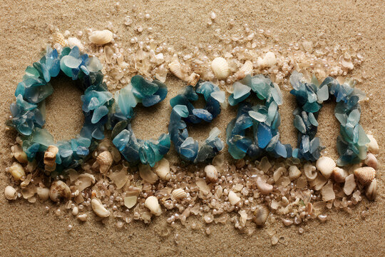 Sea Glass Ocean Typography, the word "OCEAN" is beautifully spelled out using frosted blue and teal shards of sea glass, nestled in a textured bed of tiny seashells and smooth beach sand