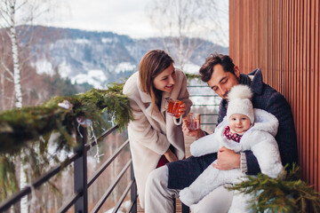 Fototapeta premium Happy family drinking tea enjoying snowy mountains landscape from hotel balcony. Mom and dad having fun with toddler baby girl