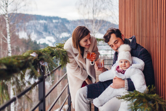 Happy family drinking tea enjoying snowy mountains landscape from hotel balcony. Mom and dad having fun with toddler baby girl