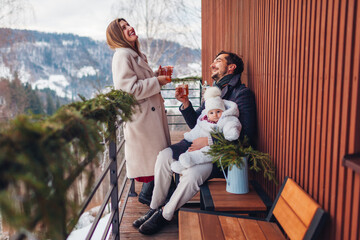 Fototapeta premium Happy family drinking tea enjoying snowy mountains landscape from hotel balcony. Mom and dad having fun with toddler baby girl