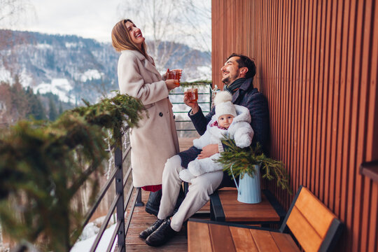 Happy family drinking tea enjoying snowy mountains landscape from hotel balcony. Mom and dad having fun with toddler baby girl