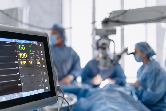 Medical monitor displays heart rate, blood pressure and blood oxygen level during surgery. Doctors work in the background.