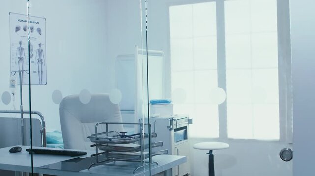 Handheld footage shows a bright empty clinic consultation room with desk, chair, document trays and examination furnishings behind a glass partition, reinforcing the empty clinical setting.