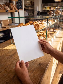 Hands holding a blank paper mockup before a sunlit bakery pastry display