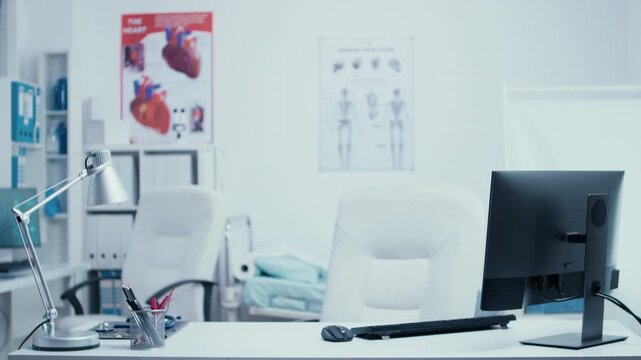 Empty medical consultation office with desk, computer, examination furnishings and anatomical posters, offering a clean clinical workspace with no people, reinforcing the empty clinical setting.