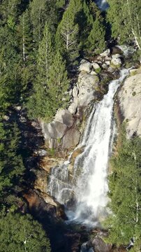 Aerial view of Cascada de Molieres waterfall cascading through alpine forest and granite mountain peaks, Val d'Aran, Pyrenees, Spain