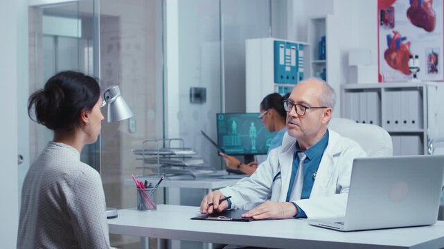 Senior male doctor listens as a woman explains her concerns during a face-to-face medical consultation in a modern clinic, reinforcing patient communication, clinical assessment and supportive care.