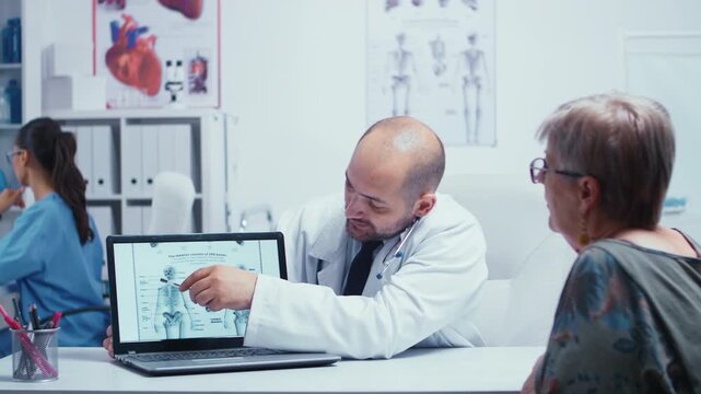 Doctor points to a skeletal anatomy chart on a laptop while explaining bone and body information to an older woman in a modern medical office.